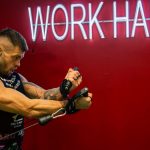 A muscular man exercising in the gym with a motivational 'Work Hard' neon sign on a red background.
