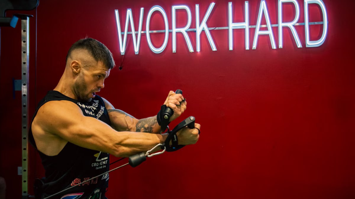 A muscular man exercising in the gym with a motivational 'Work Hard' neon sign on a red background.